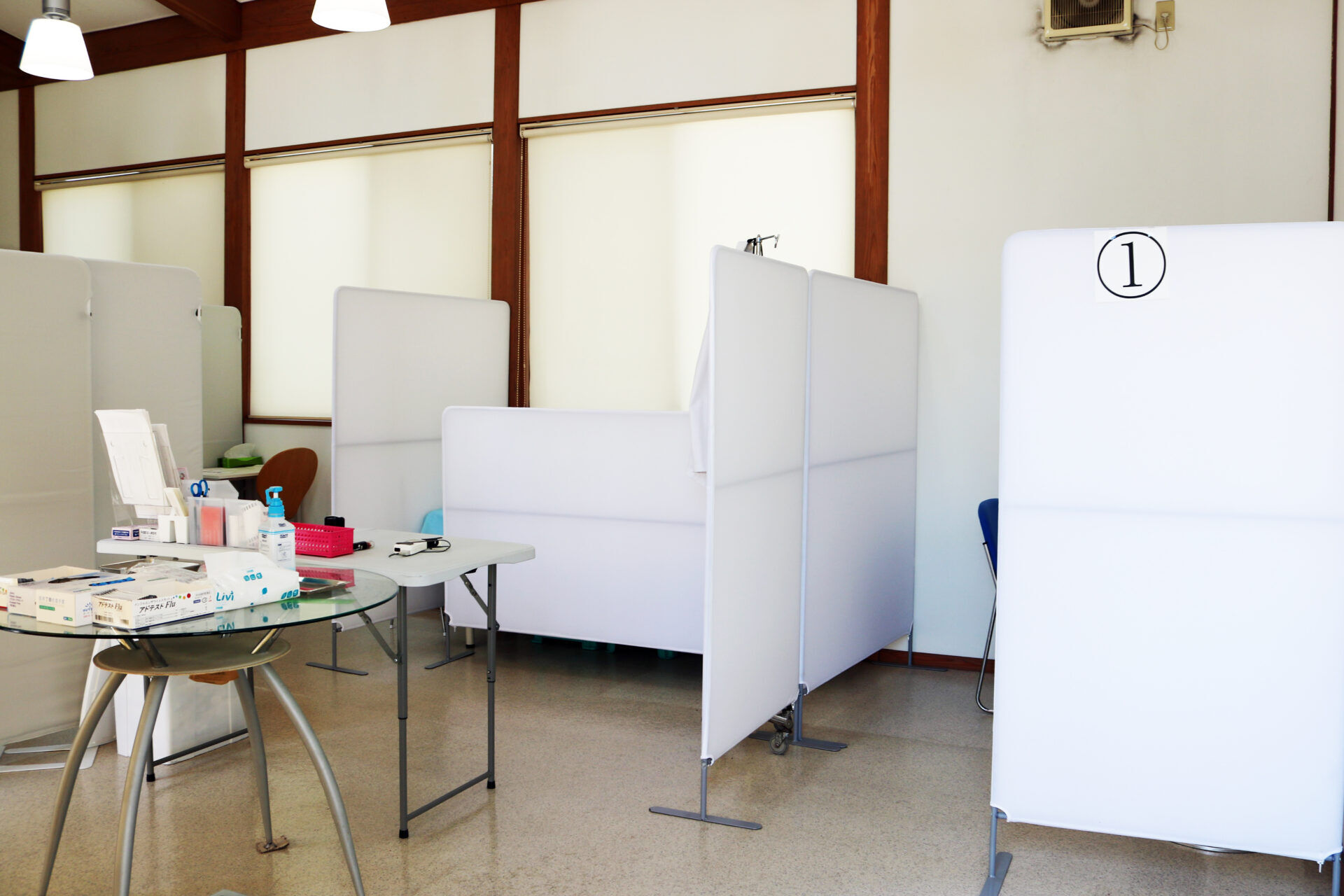 Private clinic workspace with white privacy booths, a table of medical supplies, and a hand sanitizer bottle visible.
