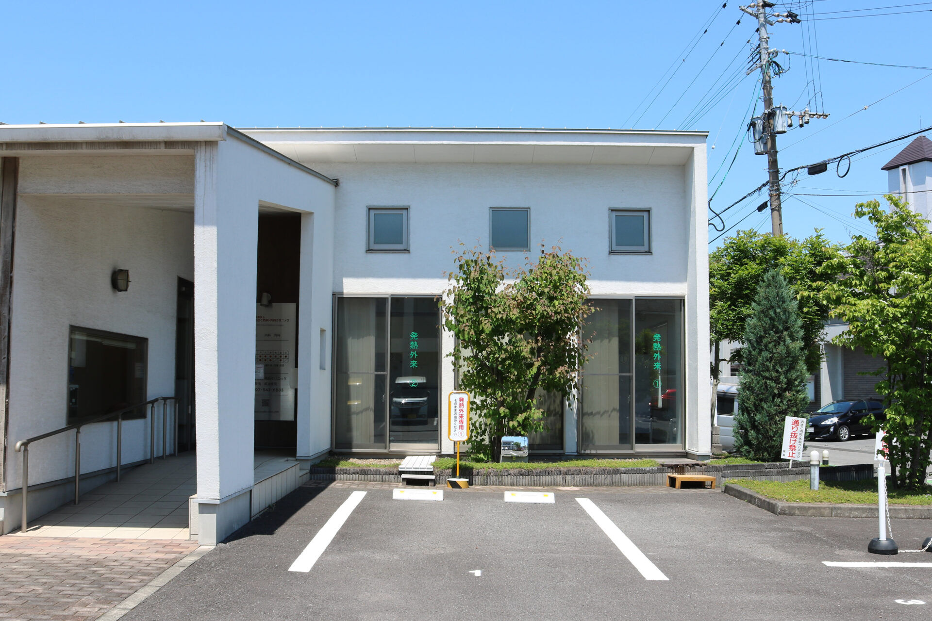 White modern building facade with glass doors, small trees, and a paved parking area in front under a blue sky