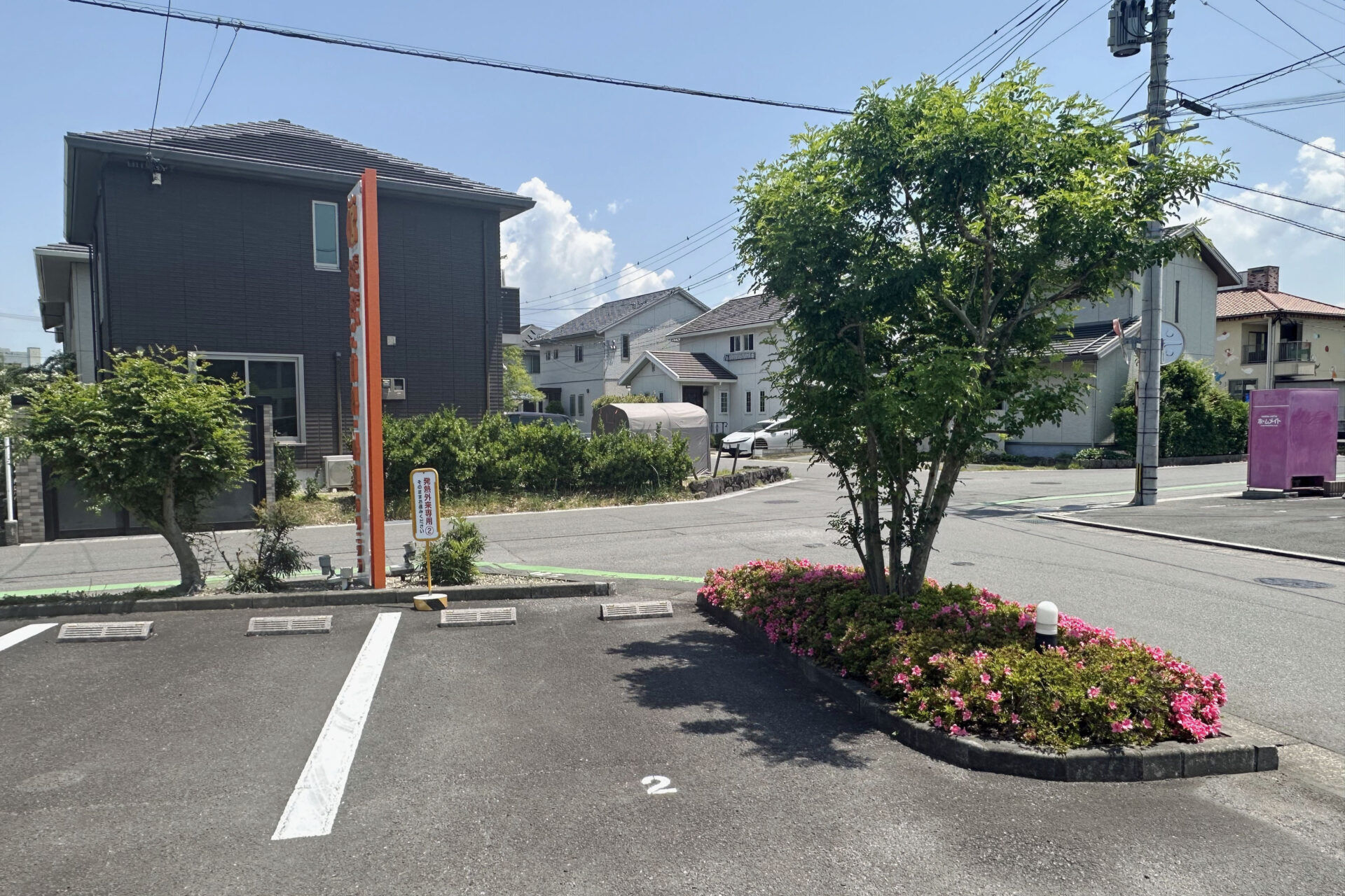 Parking lot with a marked space'2', a circular flower bed with pink blooms, and trees beside modern buildings under a blue sky.