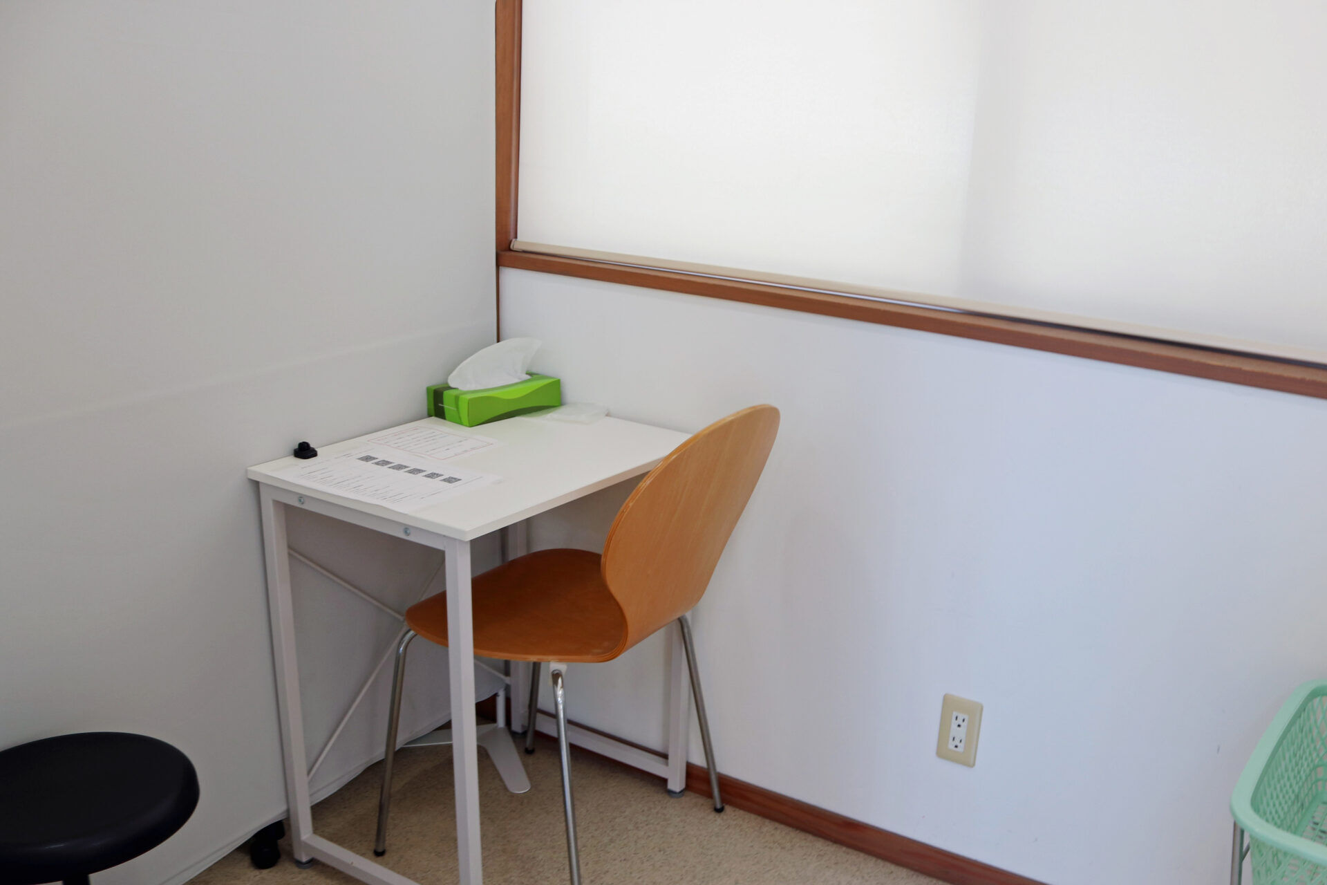 Small white desk in a corner with a green tissue box and white tissues, a wooden chair, and a black stool nearby.