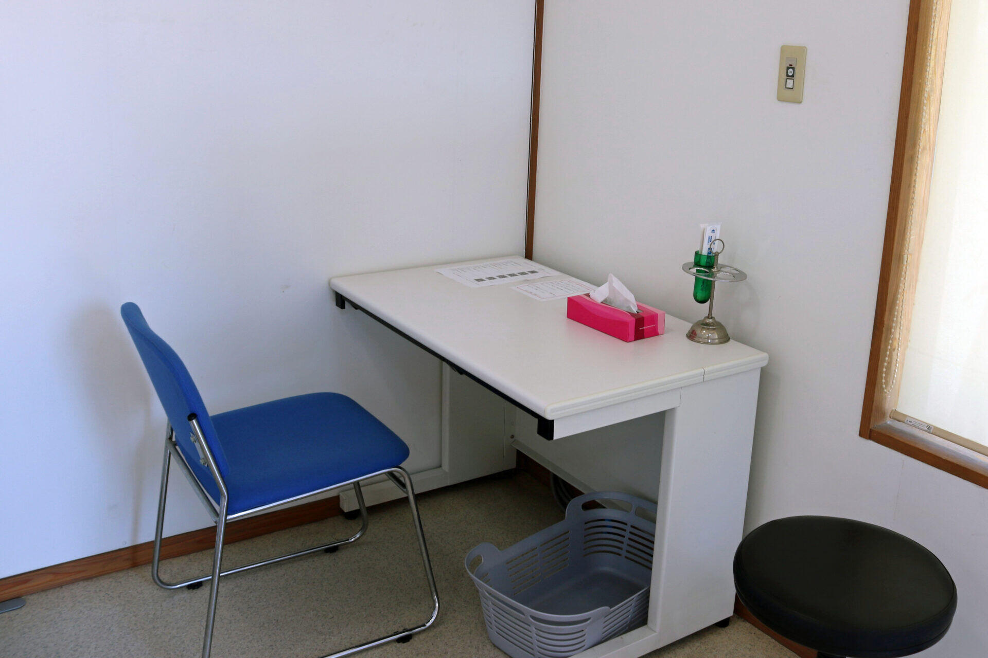 A small clinic corner with a white desk, blue chair, tissue box, and a green medical device on a stand near a window frame.